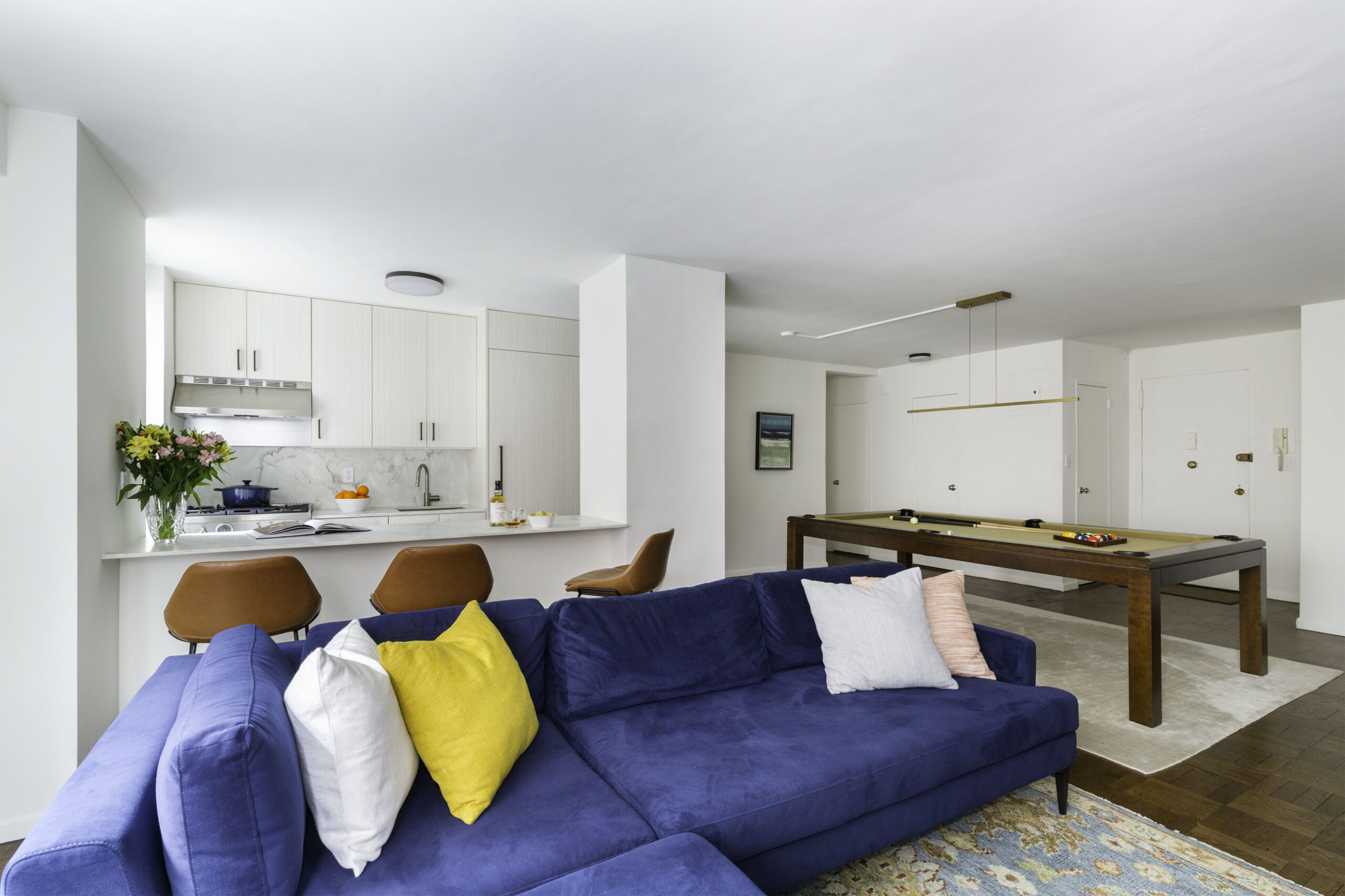Blue sofa with pillows, wood flooring and rug, with kitchen peninsula in the background and pool table at the side