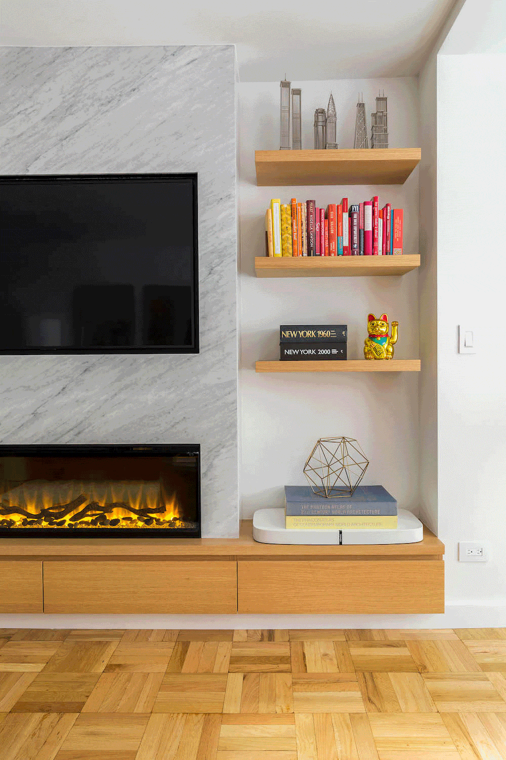 Close-up of marble-clad fireplace wall with recessed TV above linear electric fire, oak floating shelves styled with books and décor, and wood console.