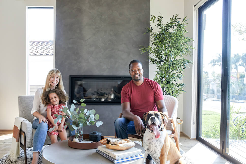 A couple with their child sitting in a modern family living room with linear fireplace wall, floor to ceiling windows, and indoor plant near glass doors.