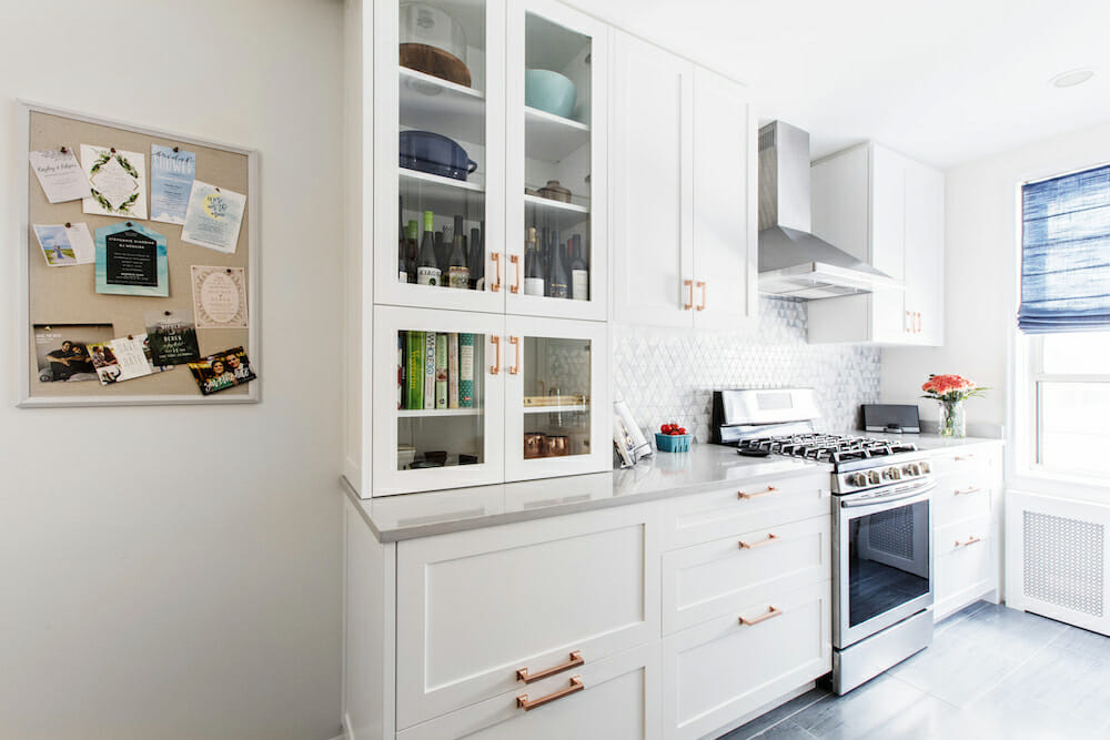 Remodeled Bronx kitchen containing storage wall with glass-front cabinets, range and hood, quartz counters, and geometric tile backsplash.